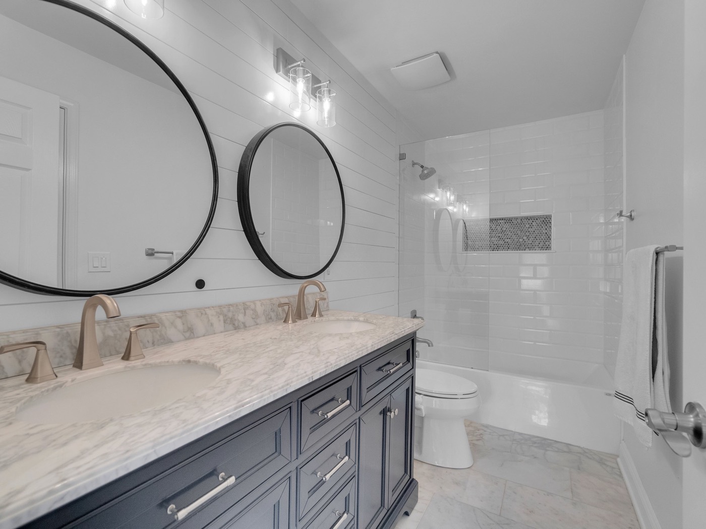 After: Navy double vanity with marble top and round black mirrors, tub with glass panel and white subway tile